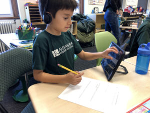 ACDS Lower School boy working at table with headphones and ipad and workbook